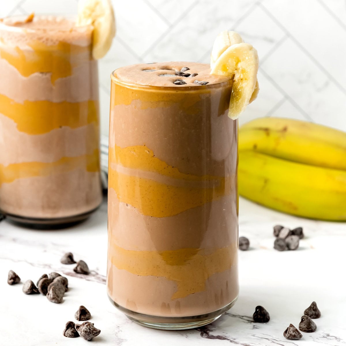 A glass of chocolate banana smoothie with banana slices and chocolate chips, set by bananas and more smoothie.