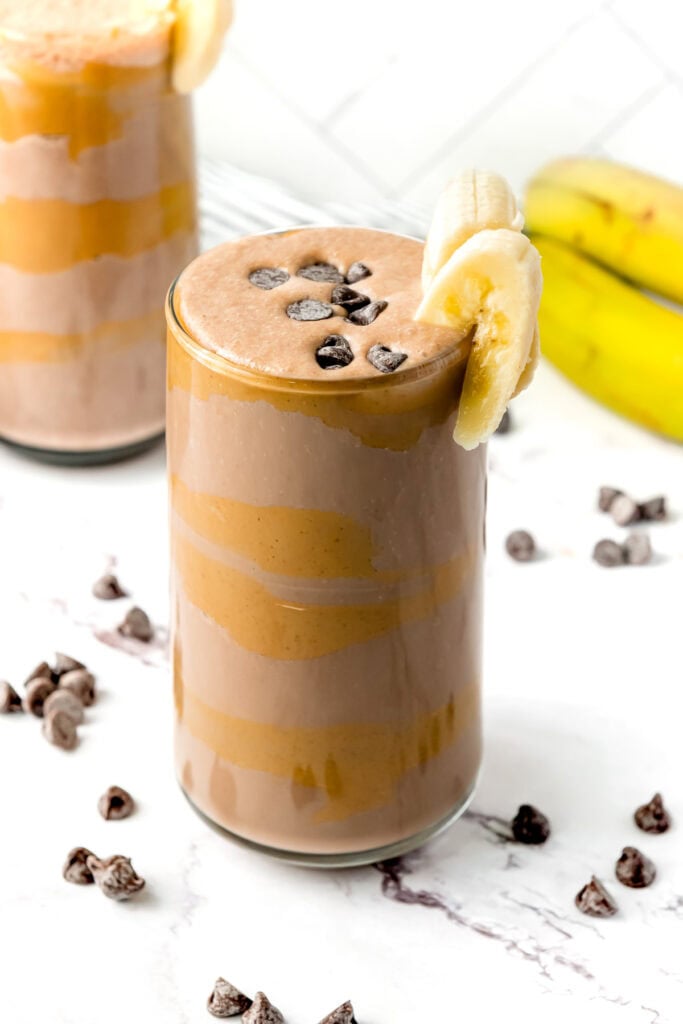 Glass of layered chocolate banana smoothie topped with chocolate chips and banana slices, with bananas in background.