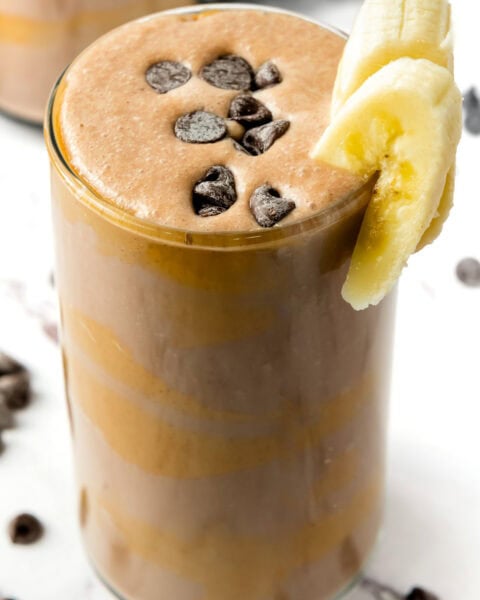 A glass of Chocolate Peanut Butter Power Smoothie topped with chocolate chips and banana slices on the rim.