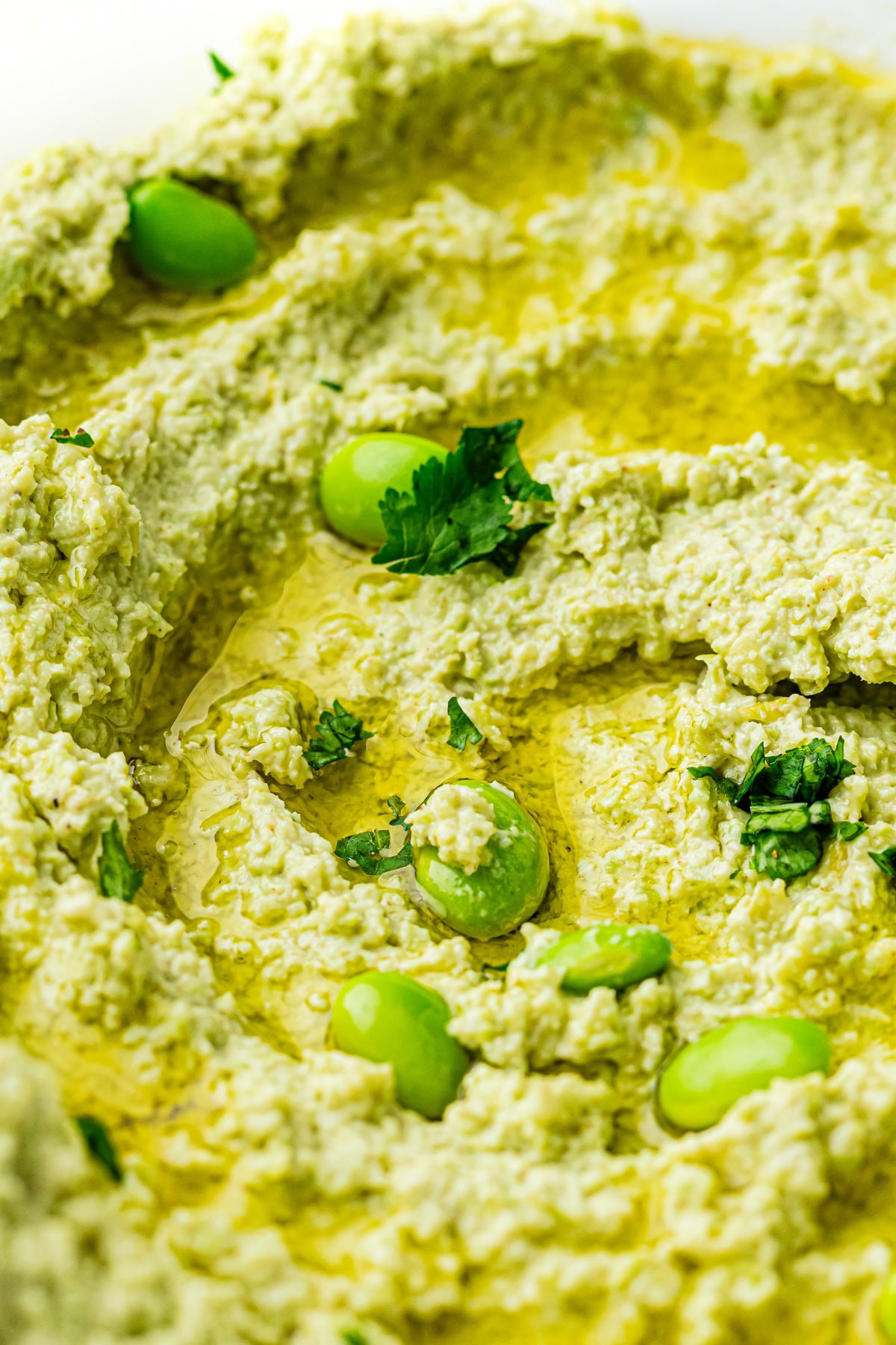 Close-up of green hummus topped with olive oil, edamame beans, and fresh herbs.