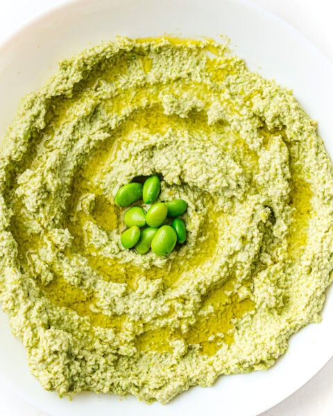 Green hummus with a swirl of olive oil and edamame beans, served on a white plate.
