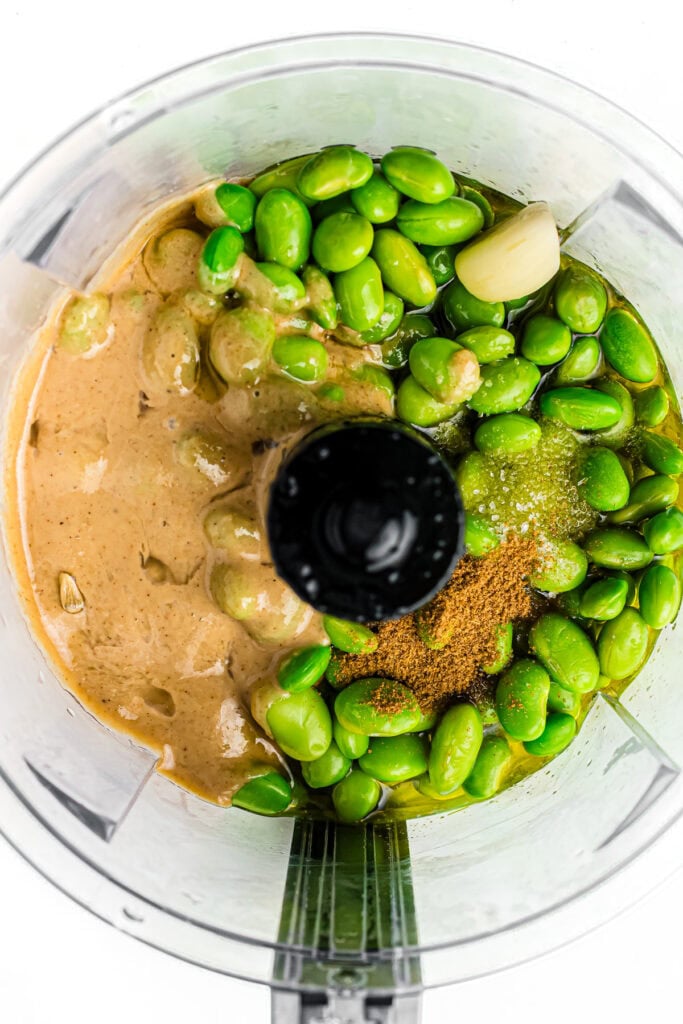 Overhead view of a food processor with edamame, garlic, seasonings, and tahini inside, ready to blend.
