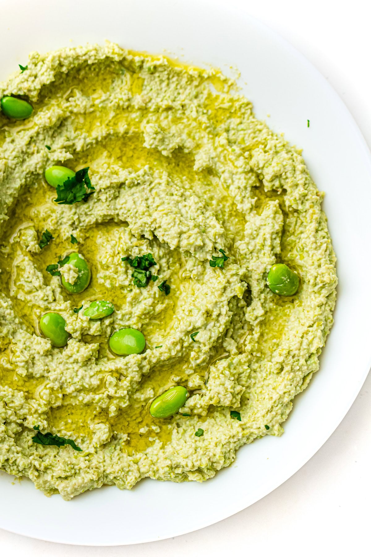 Green edamame hummus topped with whole edamame beans, herbs, and a drizzle of olive oil on a white plate.
