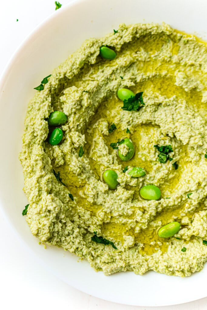 Green edamame hummus garnished with whole edamame beans and herbs on a white plate.