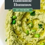 A bowl of green edamame hummus garnished with whole edamame beans and herbs.