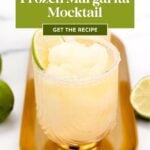 A frozen margarita mocktail with a lime wedge in a glass, on a gold tray, surrounded by limes.
