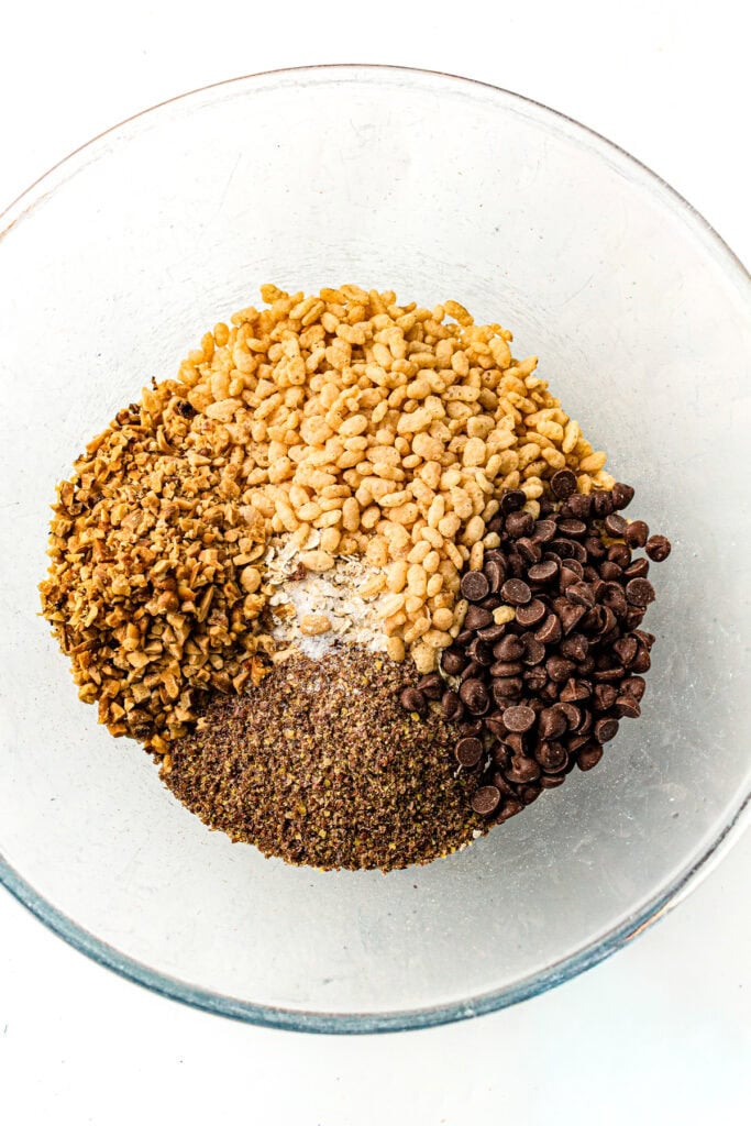A glass bowl with crispy rice, mini chocolate chips, chopped nuts, and ground flaxseed, viewed from above.