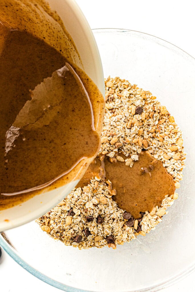 Almond butter mixture being poured into a bowl of oats, chocolate chips, and seeds for baking.
