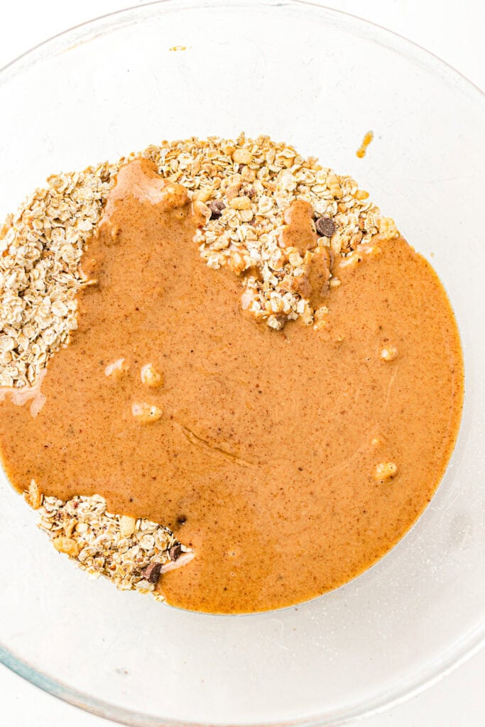 A glass bowl with oats and a creamy brown mixture, partially combined, on a white background.