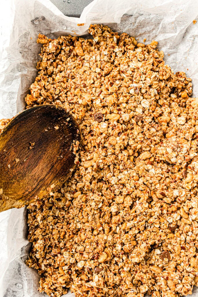 Granola mixture with oats and nuts on parchment paper, stirred with a wooden spoon.
