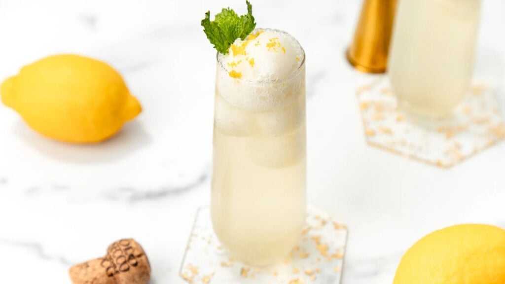 A tall glass of Lemonade Sorbet Float cocktail with ice, foam, and mint, surrounded by lemons on a white surface.