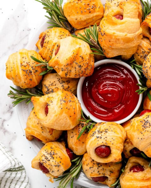 Golden pigs in a blanket with poppy seeds, garnished with rosemary, served with ketchup in a white dish.