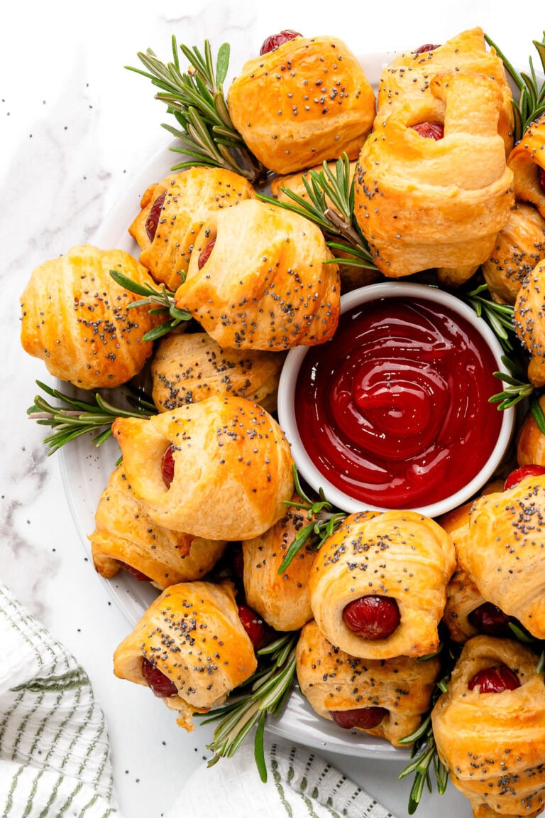 Golden pigs in a blanket with poppy seeds, garnished with rosemary, served with ketchup in a white dish.