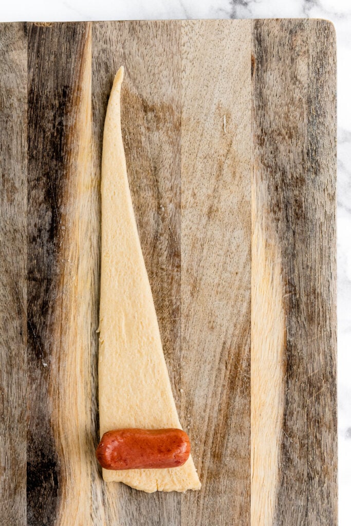 Triangle crescent roll dough with a sausage at the base on a wooden board, ready to be rolled up.