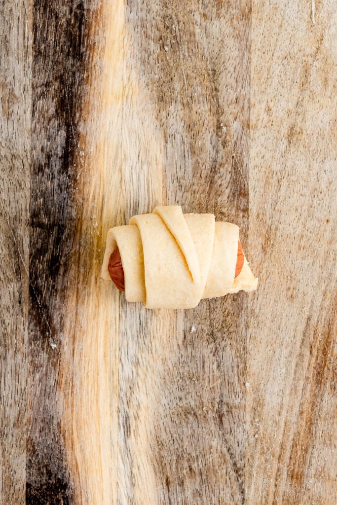 A mini sausage wrapped in dough on a wooden surface, ready to be baked.