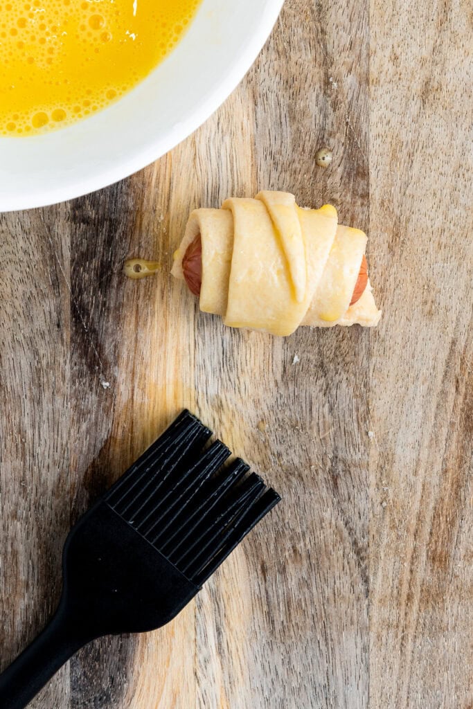 Unbaked pig in a blanket next to a bowl of beaten egg and a black pastry brush on a wooden surface.