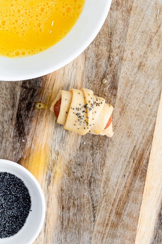 Uncooked pig in a blanket with poppy seeds on dough, beside bowls of beaten egg and poppy seeds on wood.