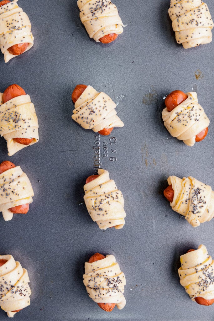 Unbaked pigs in a blanket topped with poppy seeds arranged on a dark baking sheet.