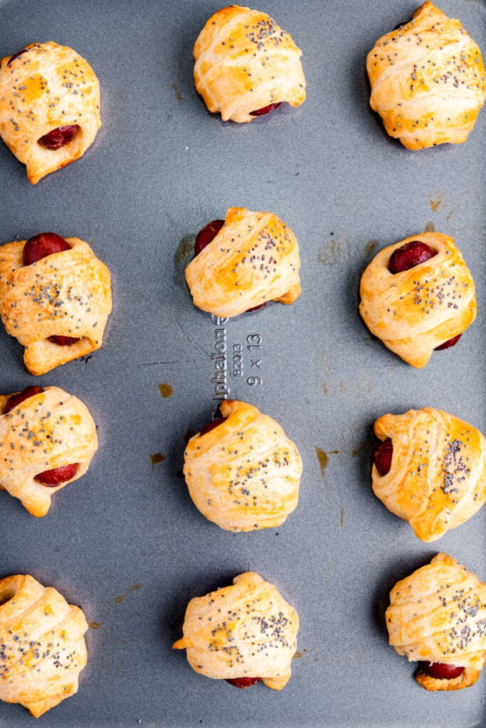 Baked pigs in a blanket on a gray baking tray, topped with poppy seeds and spaced evenly apart.