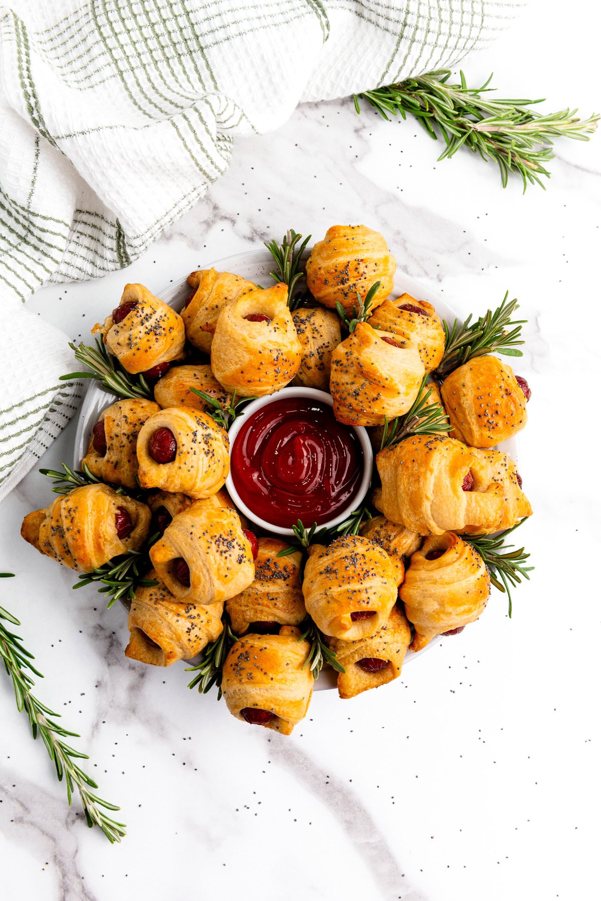 Mini crescent roll pigs in a blanket arranged in a circle with rosemary and ketchup on a marble surface.