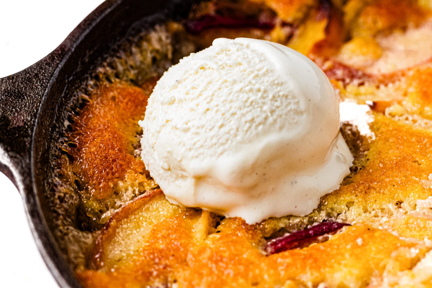 A scoop of vanilla ice cream melting on top of a golden peach cobbler in a cast iron skillet.