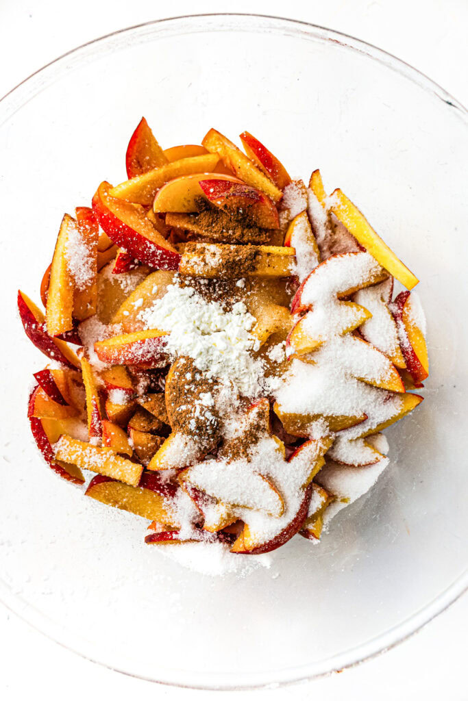 Sliced nectarines in a bowl topped with sugar, cinnamon, and cornstarch, ready to be mixed.