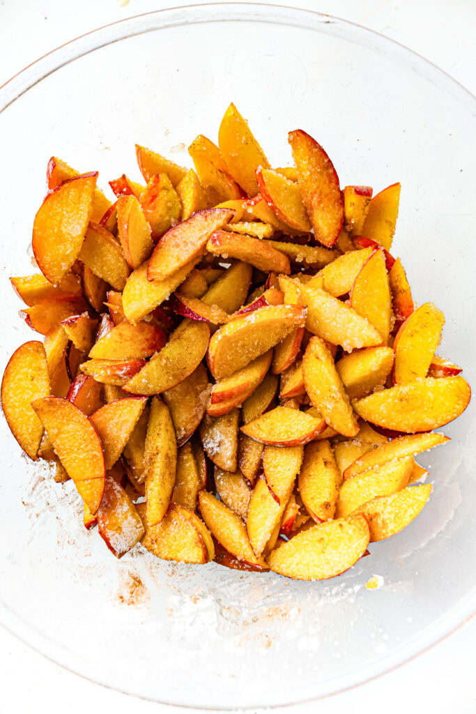 Sliced peaches in a clear bowl coated with sugar and spices, ready for baking or cooking.