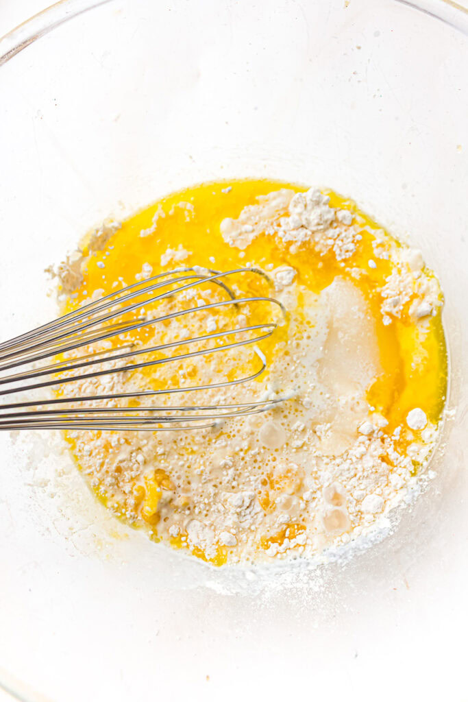 A metal whisk in a bowl with flour, eggs, and liquid being mixed for baking.