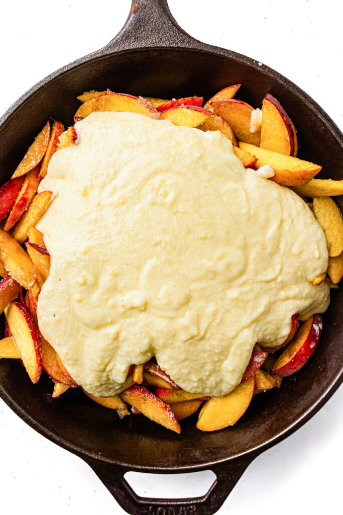 Sliced peaches in a cast iron skillet topped with an uncooked biscuit-like batter, ready for baking.