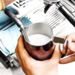 Hands steaming milk in a metal pitcher under a coffee machine steam wand.