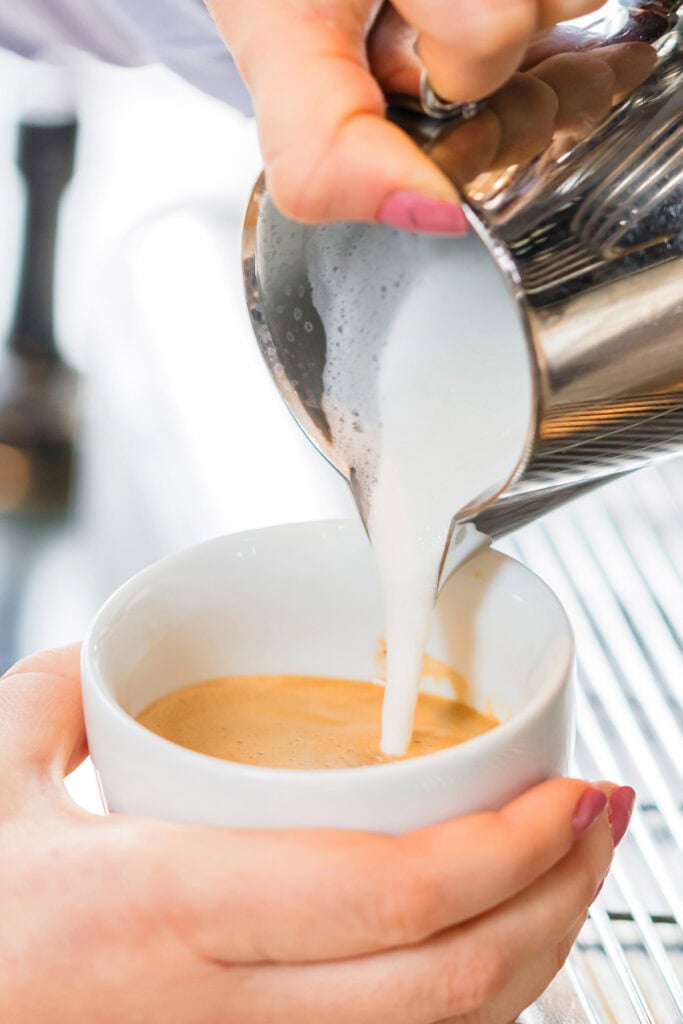Close-up of milk being poured from a metal pitcher into a cup of coffee, held by a person with pink nails.