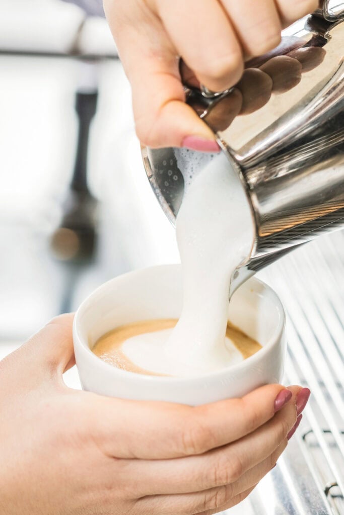 Close-up of steamed milk being poured from a metal pitcher into a cup of coffee held by a person with pink nails.