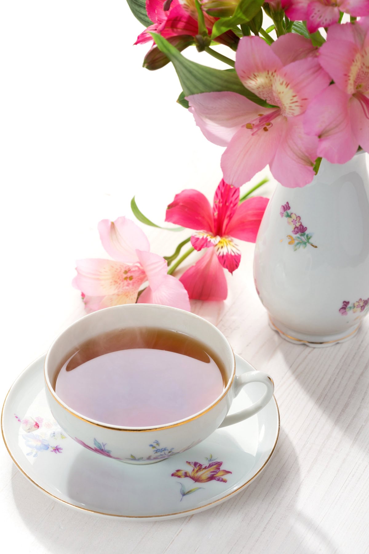 A floral teacup filled with tea sits beside a vase of pink flowers on a white surface.