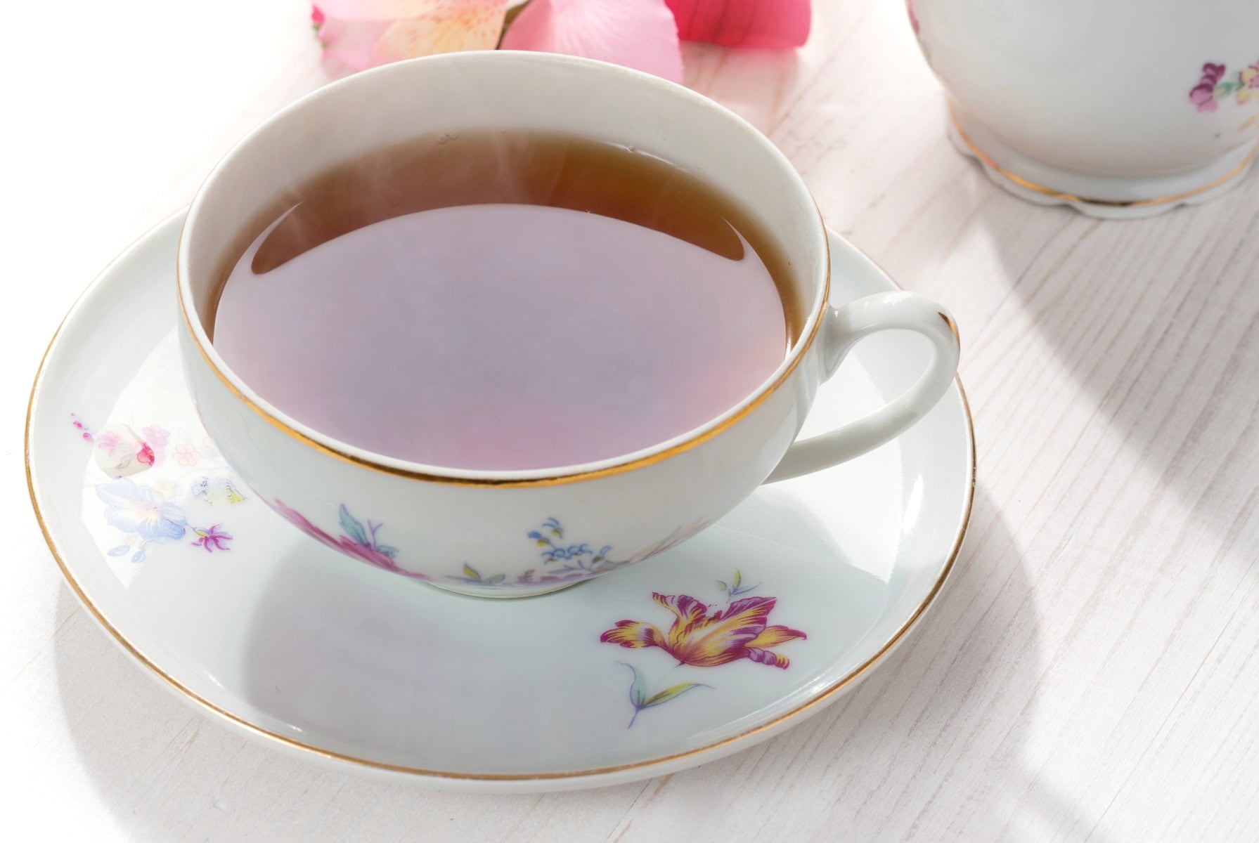 A steaming cup of tea in a floral china cup and saucer, on a white table with pink flowers in the background.
