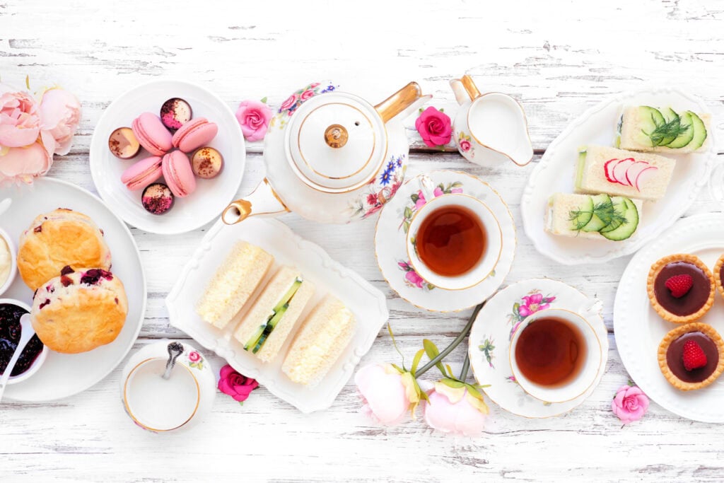 Tea set with floral china, sandwiches, scones, macarons, and tarts arranged on a white wooden table.