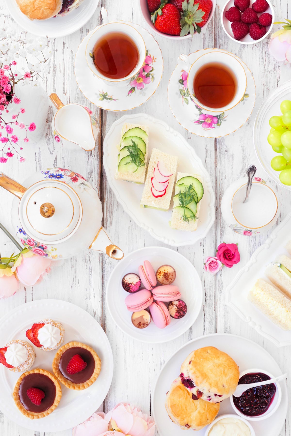 Afternoon tea spread with tea, sandwiches, scones, macarons, pastries, fruit, and floral china on white wood.