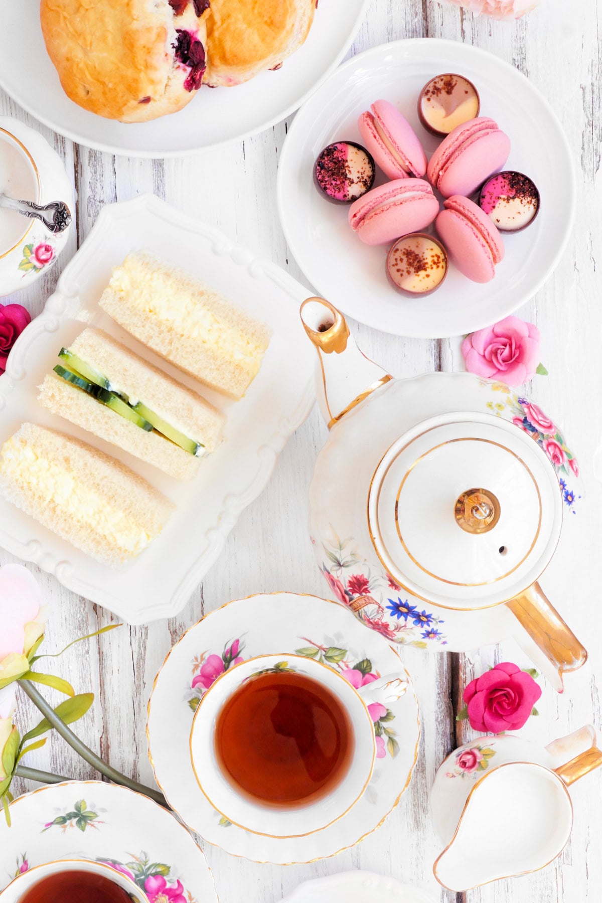 English tea set with tea, a teapot, sandwiches, macarons, pastries, and flowers on a white wooden table.