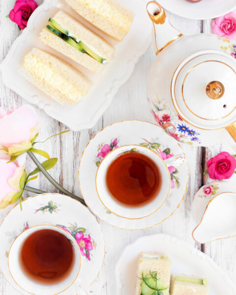 Top view of tea cups, floral china, sandwiches, macarons, and pink flowers on a white wooden table.