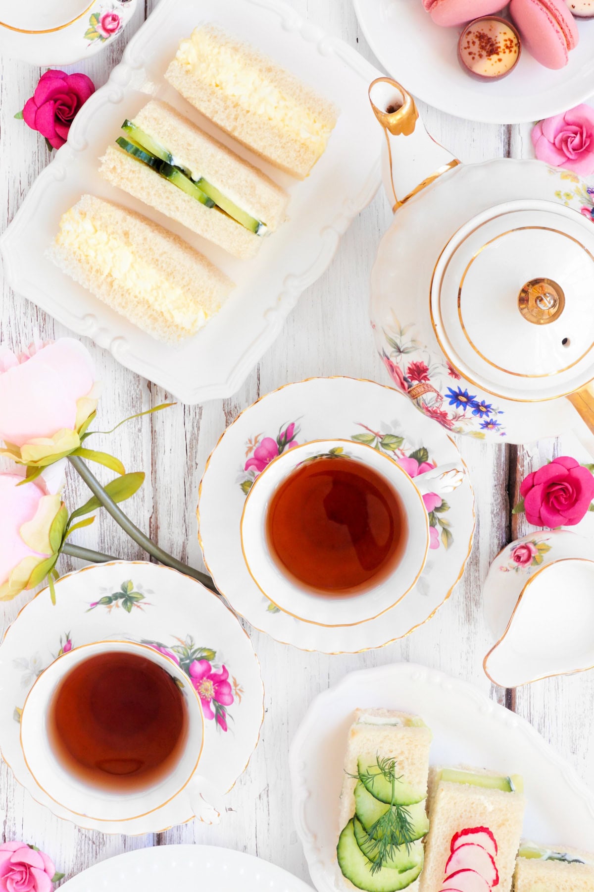 Top view of tea cups, floral china, sandwiches, macarons, and pink flowers on a white wooden table.