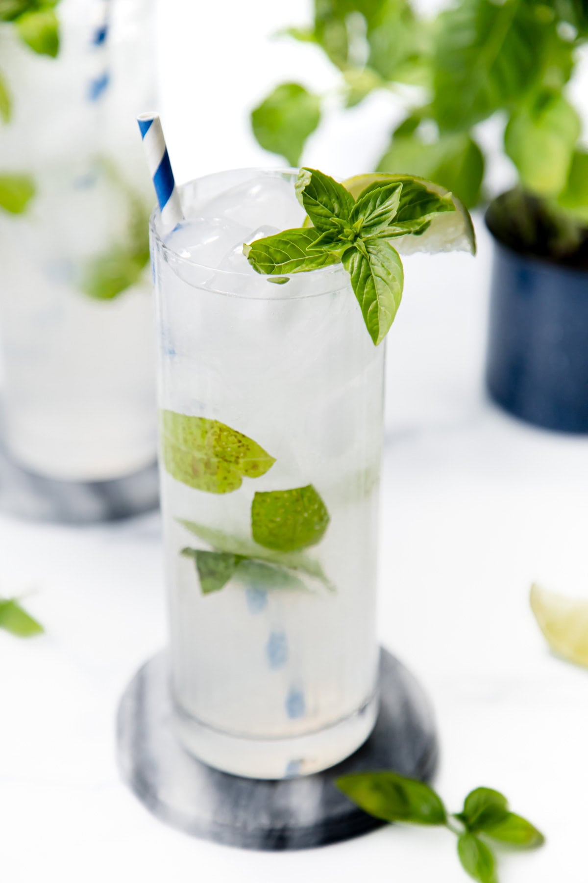 Tall glass with ice, lime, mint, and a striped straw, on a coaster, with mint leaves in the background.