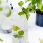 A tall glass of iced mojito with mint leaves, a lime wedge, and a striped straw on a white surface.