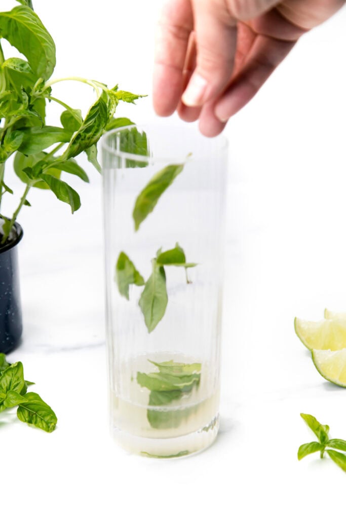 A hand drops fresh basil leaves into a tall glass with lime and basil nearby on a white surface.