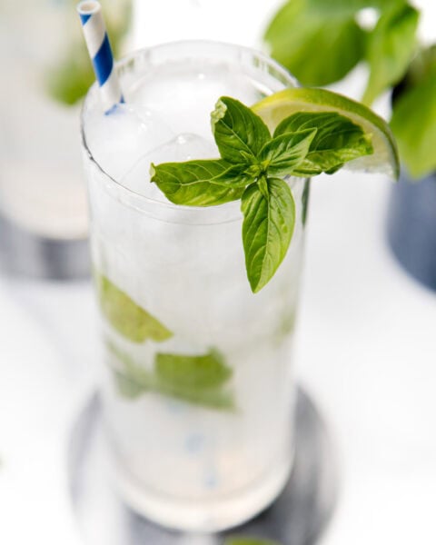 A glass of ice water with basil leaves, a lime wedge, and a blue-striped straw on a coaster.