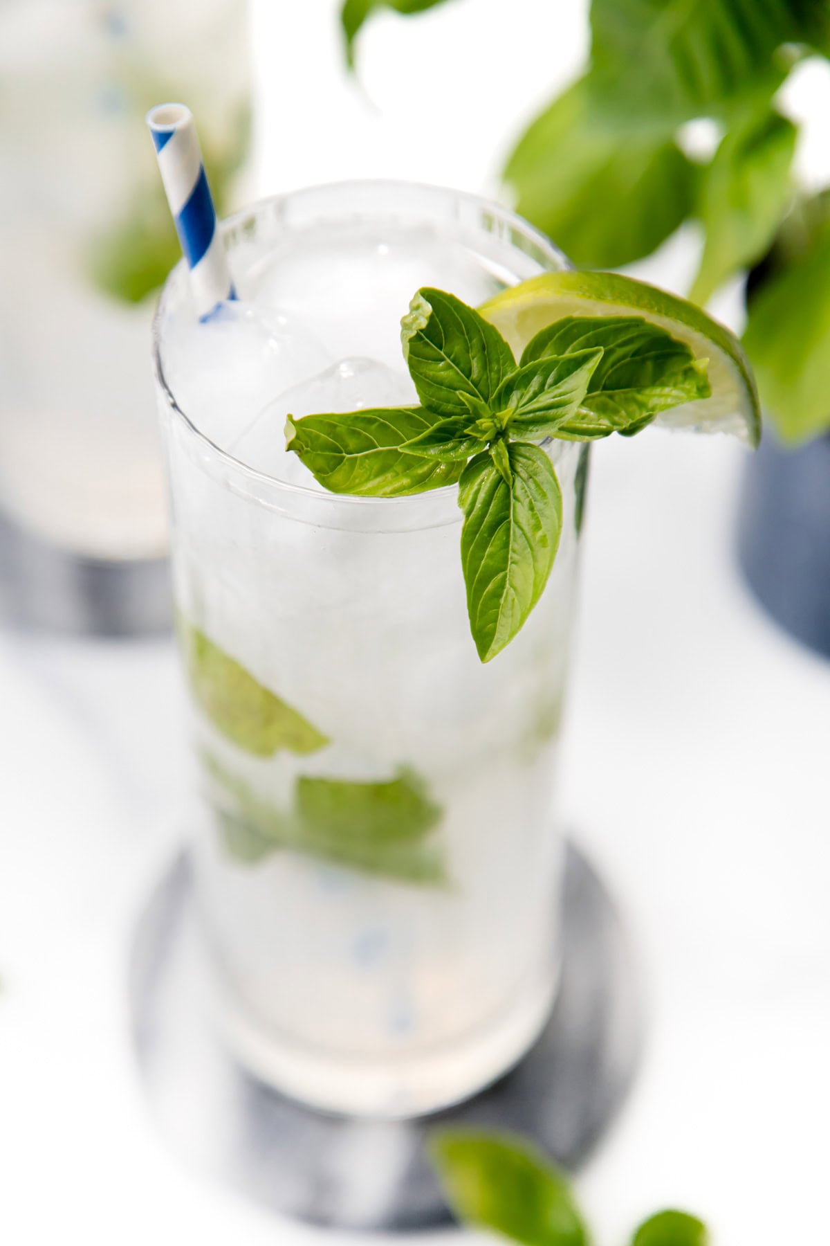 A glass of ice water with basil leaves, a lime wedge, and a blue-striped straw on a coaster.