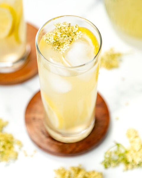 A tall glass of iced lemonade with lemon slices and a small elderflower garnish on top, on a wooden coaster.