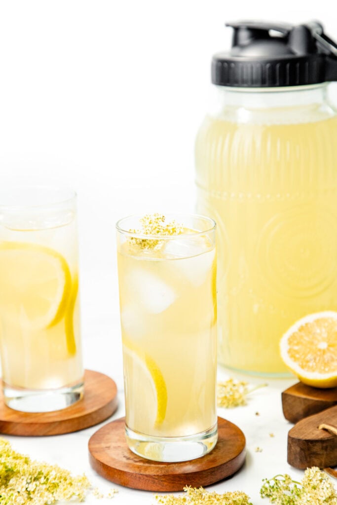 Two glasses of lemonade with lemon slices and ice, a jug of lemonade, and lemon halves on the table.