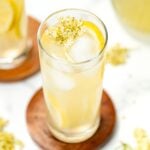 A tall glass of lemonade with ice, lemon slices, and a sprig of flowers, on a wooden coaster.