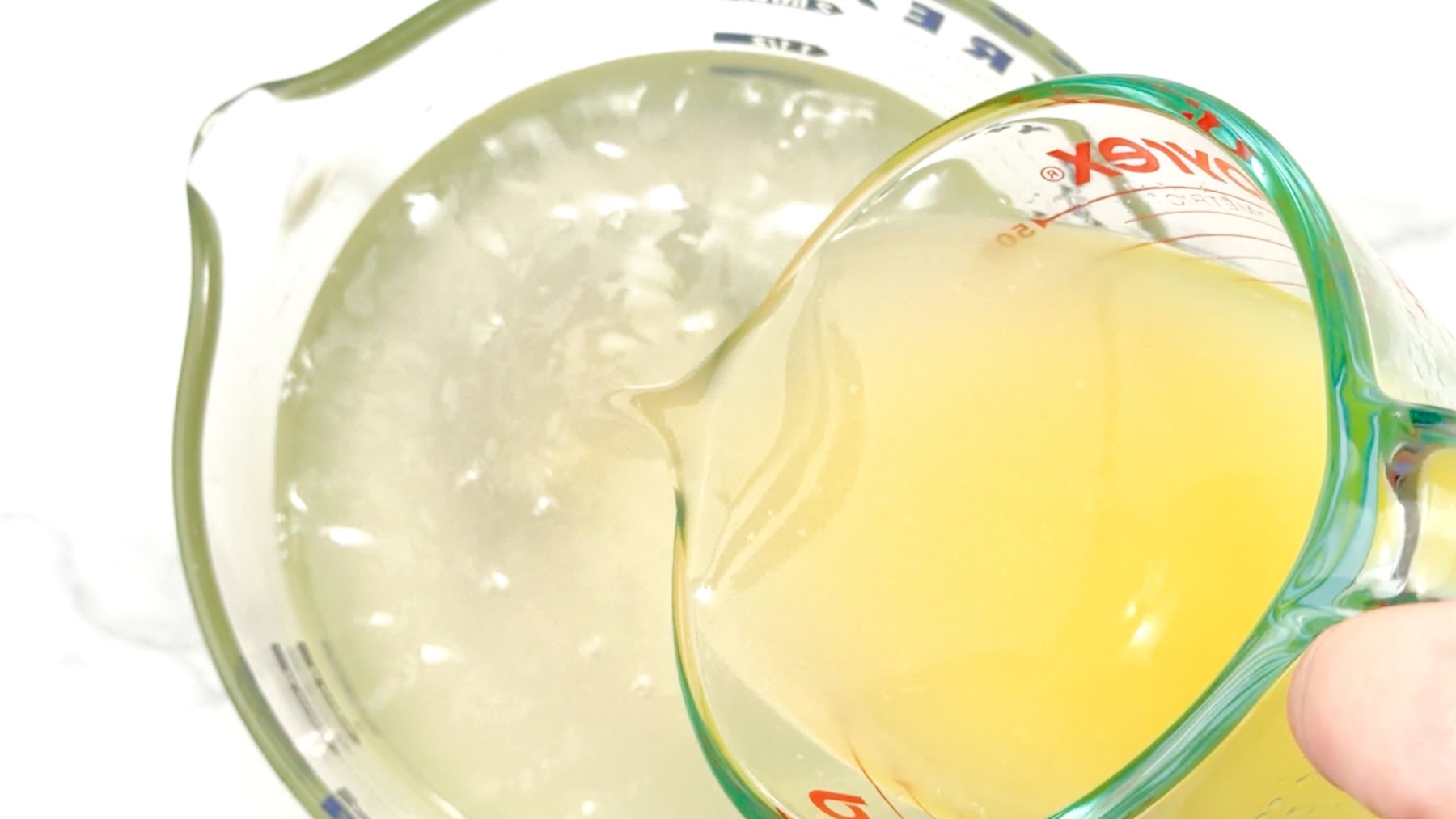 Yellow lemon juice is being poured from a glass measuring cup into another liquid in a larger measuring cup, creating the perfect base for refreshing elderflower lemonade.