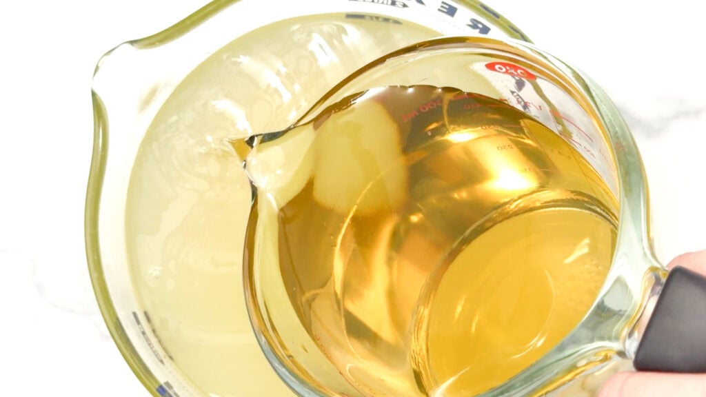 A glass measuring cup pours golden Elderflower Lemonade into another cup with a pale yellow liquid, both on a white surface.