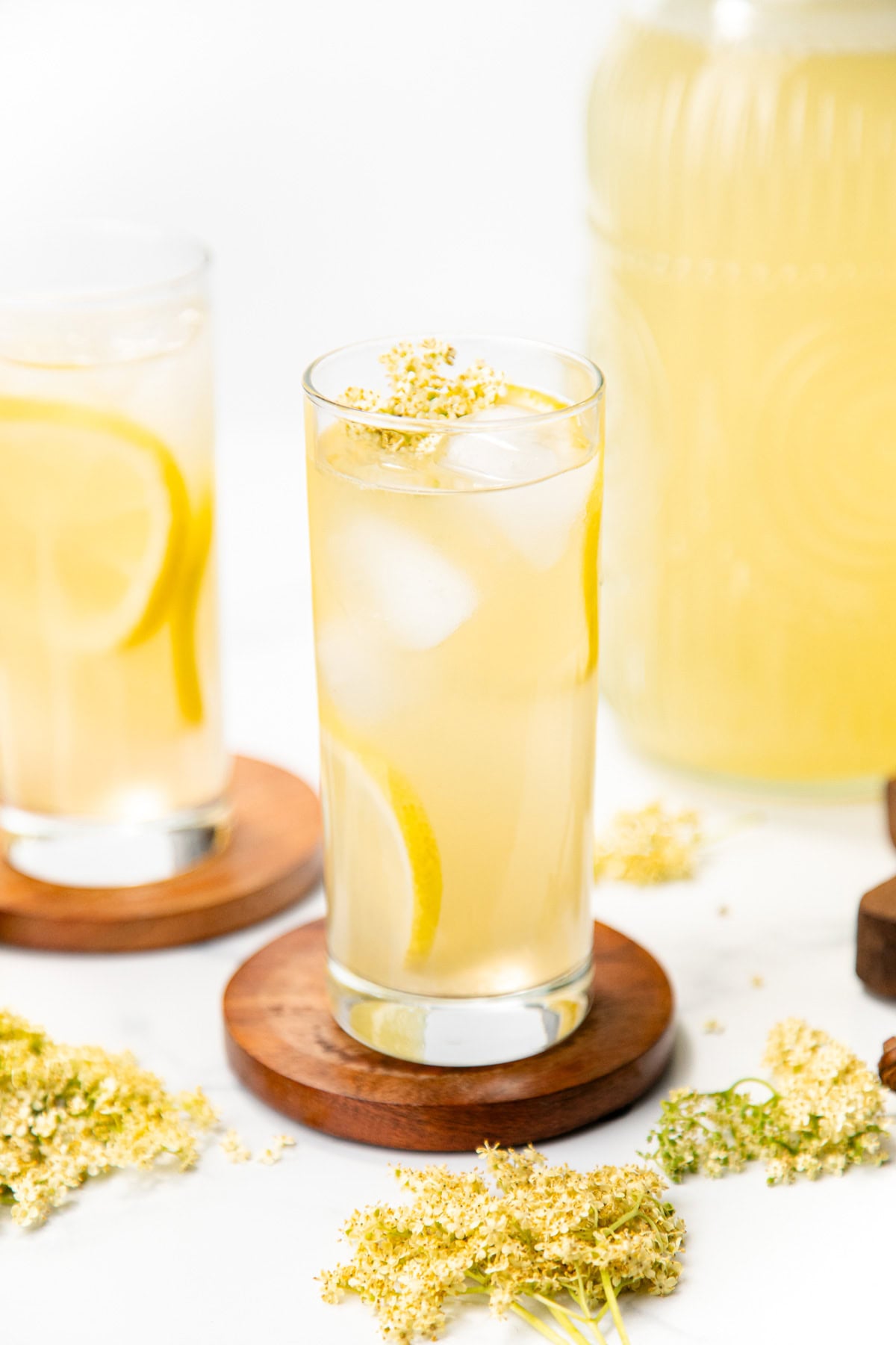 A glass of lemonade with ice, lemon slices, and flower garnish on a wooden coaster, with flowers nearby.